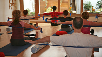 People in a Pilates Mat class sitting on mats, holding resistance bands, with arms extended.