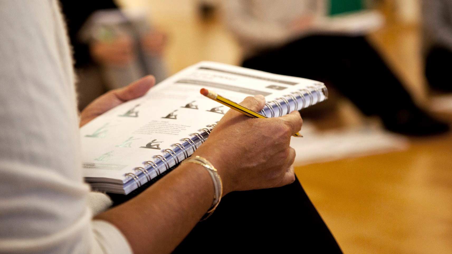 Person holding a notebook and pencil, with pages open showing text and diagrams, in a sitting position indoors.