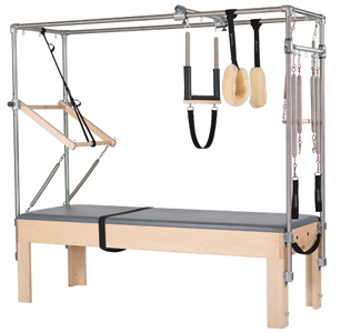 Pilates equipment Cadillac/tower, featuring a wooden base with a padded surface, metal frame, pulleys, straps, and springs for exercise and stretching.