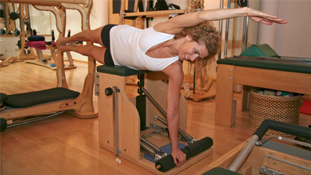 Kelly Mckinnon performing a Pilates exercise on a chair in a Pilates studio, extending one arm and leg while maintaining balance.